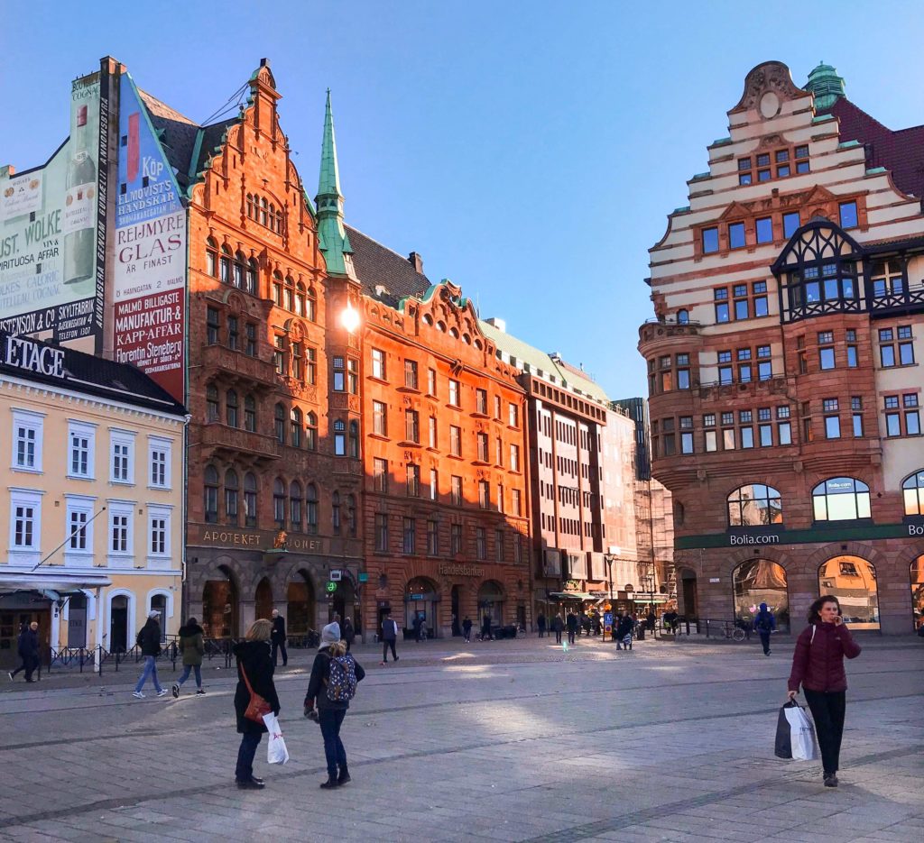 Bordo renkli Amsterdam evlerine benzeyen bir mimari tasarım ile inşa edilmiş binalar, yürüyerek geçen insanların yer aldığı bir fotoğraf karesi.