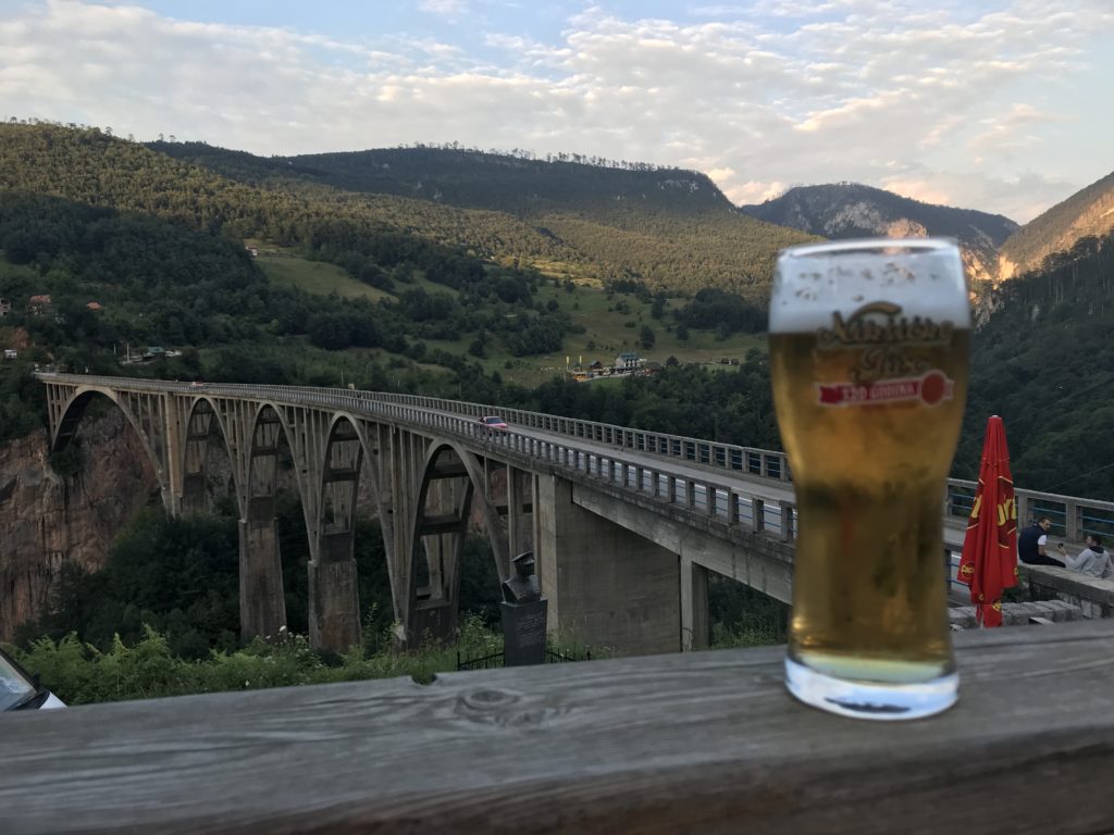 Tahta bir zenim üzerine yerleştirilmiş bira bardağının köprü ve yemyeşil bir doğa manzarası ile çekilmiş görüntüsü.