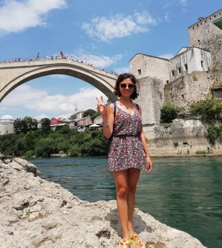 Mostar Köprüsnün altında nehir kenarında gülümseyip poz veren gezginstar ın görüntüsü.