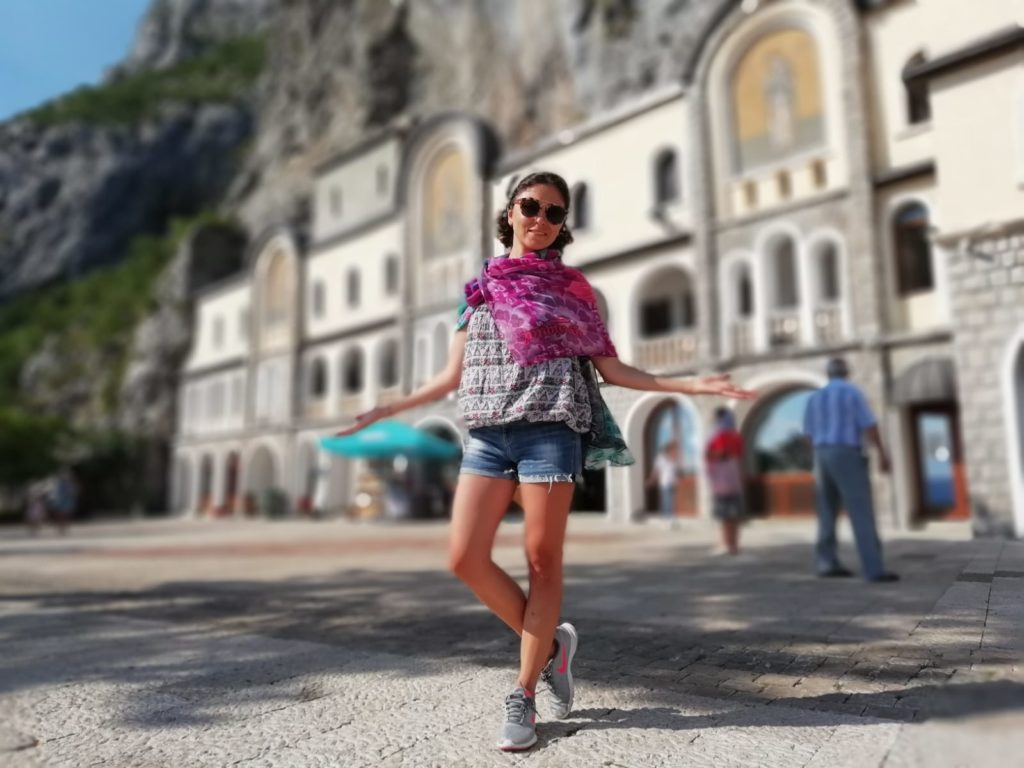 Ostrog manastırı gezi rehberi yazısında ellerinde iki yana açmış manastırın önünde poz veren kadın.