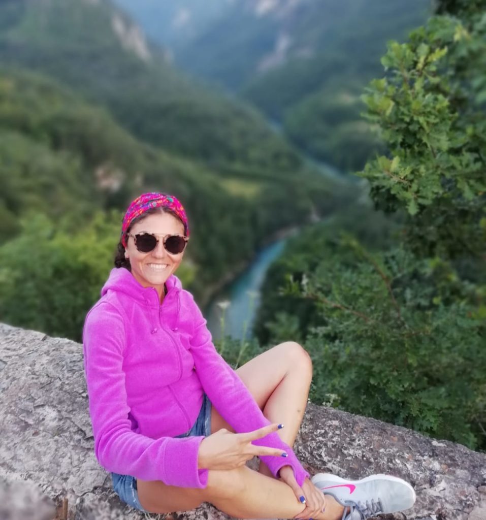 Duvar üzerine oturmuş gülümseyip poz veren gezginstar, arkada yemyeşil bir doğa ve nehrin yer aldığı gezi fotoğrafından bir kare.