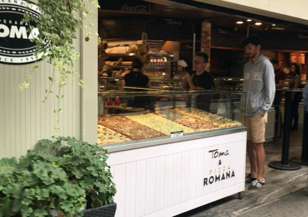 Beyaz renkli pizza dükkanının önünde sipariş alan garsonlar ve çeşitli pizzaların olduğu bir tezgahın Belgread'da ne yenir ? yazısından görüntüsü.