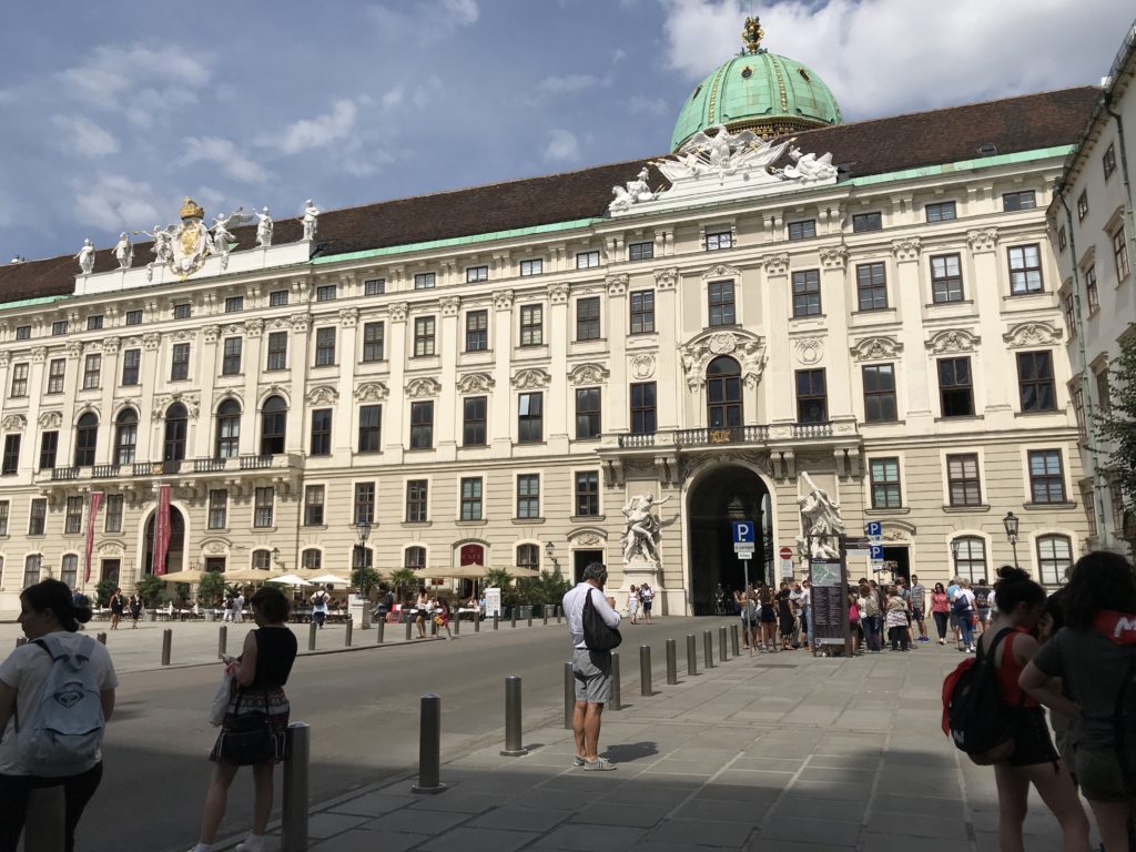 Krem rengi tarihi bir saray, yeşil renkte bir kubbe ve etrafı meraklı gözlerle inceleyen turistlerden oluşan bir gezi fotoğrafı.