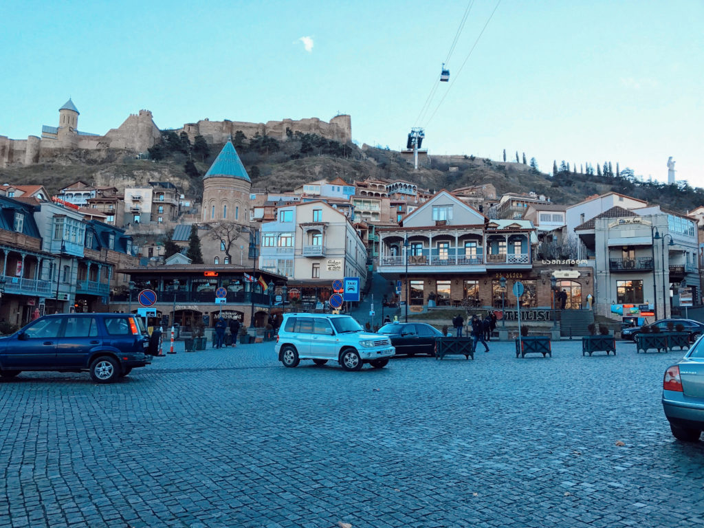 Tepe üzerinde kale surları, tepenin yamacındaki evler ve arabaların olduğu bir şehir görüntüsü.