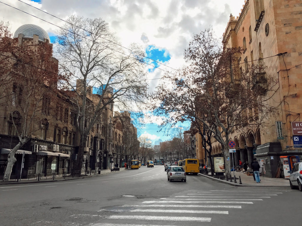 Geniş bir cadde üzerindeki binalar , araçlar ve ağaçlarında yer aldığı bir şehir görüntüsü.