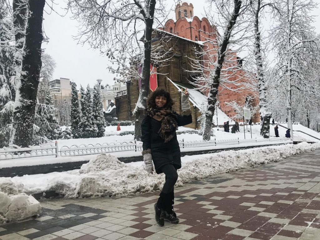 Karlarla kaplı bir park, parkın içerisinde pembe renkte ahşaplarında kullanılarak inşa edilmiş bir bina önünde poz veren bir kadın görüntüsü.