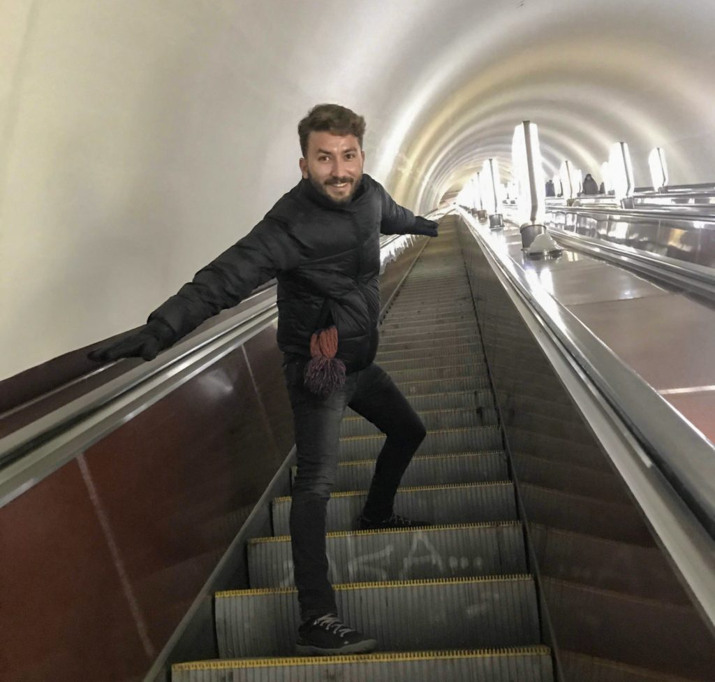 Kiev metrosunda yürüyen merdivenlerde ellerini iki yana açarak poz veren bir erkeğin görüntüsü.