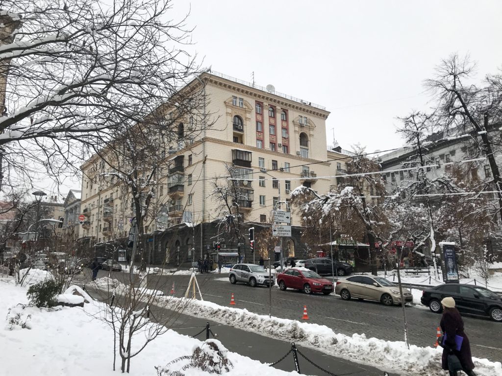 Karlarla kaplı kaldırım, buz tutmuş ağaçlar ve krem rengi yüksek katlı apartmanın yer aldığı görüntü.