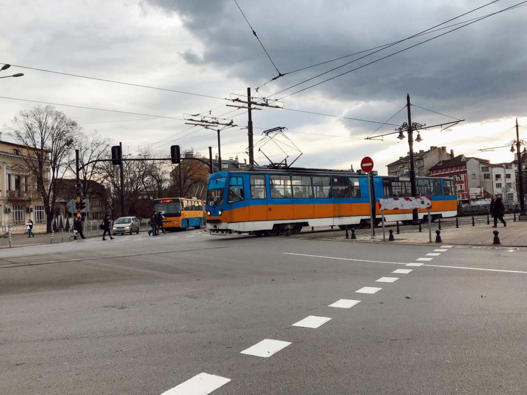 Şehirde toplu taşıma için kullanılan sarı ve mavi renkte bir tramvay ve karşıdan karşıya geçen insaların yer aldığı Sofya ulaşım yazısından bir görüntü.