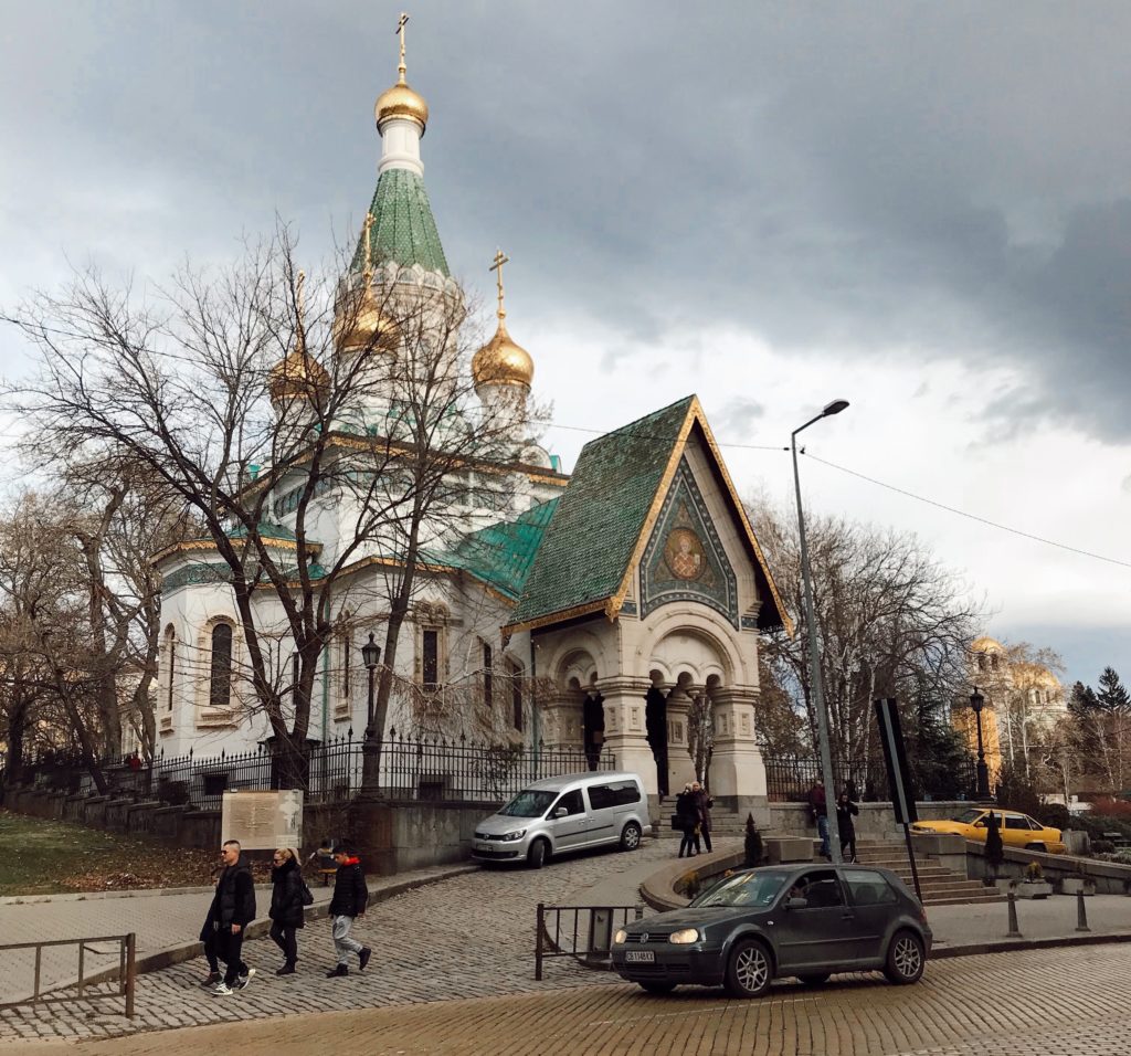Yeşil çatısı, altın varaklı işlemeleri ile cadde üzerinde yer alan bir kilise ve hemen önünde yürüyen insanların yer aldığı fotoğraf.
