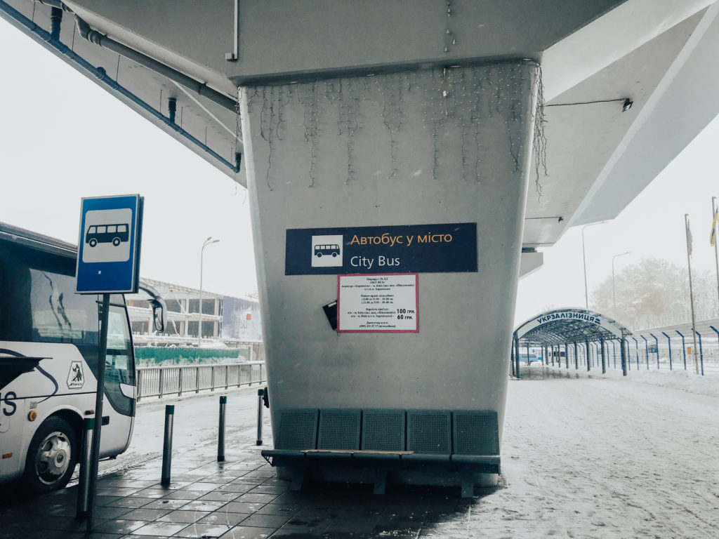 Köprü altında bekleyen gri renkte otobüs ve nereden nereye gittiğini ile fiyatın yazılı olduğu tabelaların yer aldığı görüntü.