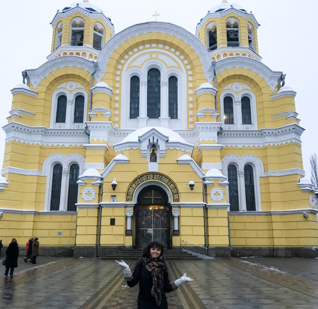 Sarı ve beyaz rebklerde boyanmış katedral önünde ellerini iki yana açarak poz veren bir kadın görüntüsü.
