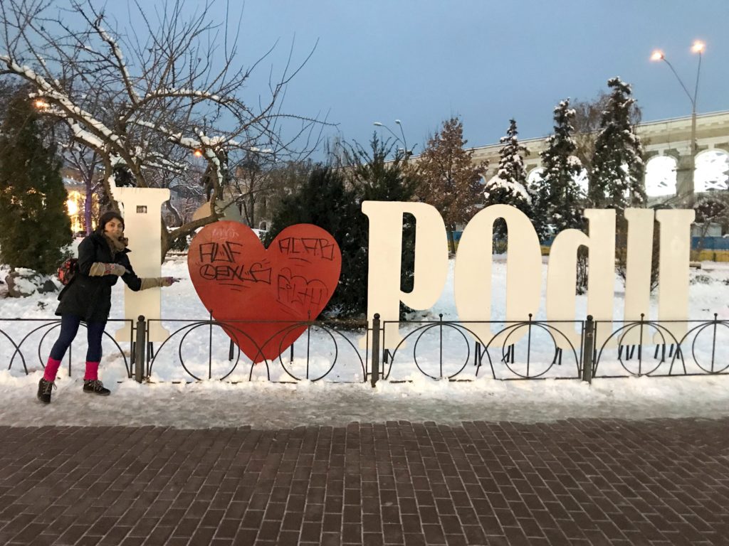 Karlarla kaplı bir parkta, l love Podil yazısı önünde poz veren bir kadın.