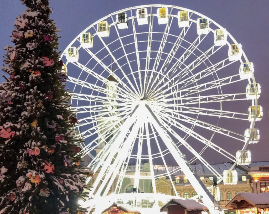 Beyaz ışıklarla aydınlatılmış dönme dolap, hemen önünde yükselen kocaman bir noel ağacının yer aldığı görüntü.