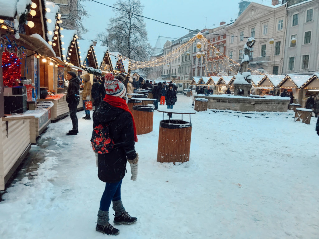 Kırmızı çantası, kırmızı atkısı ve beyaz beresi ile karlı bir günde noel pazarında ahşap kulübelerin önündeçekilmiş görüntü.