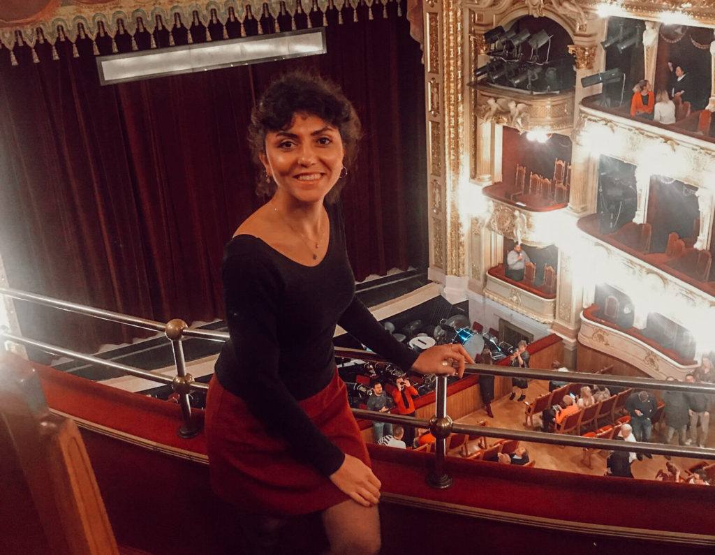 Opera salonunda, arkasında bordo renkte bir perrde, balkon korkuluklarına tutunarak poz veren bir kadının görüntüsü.