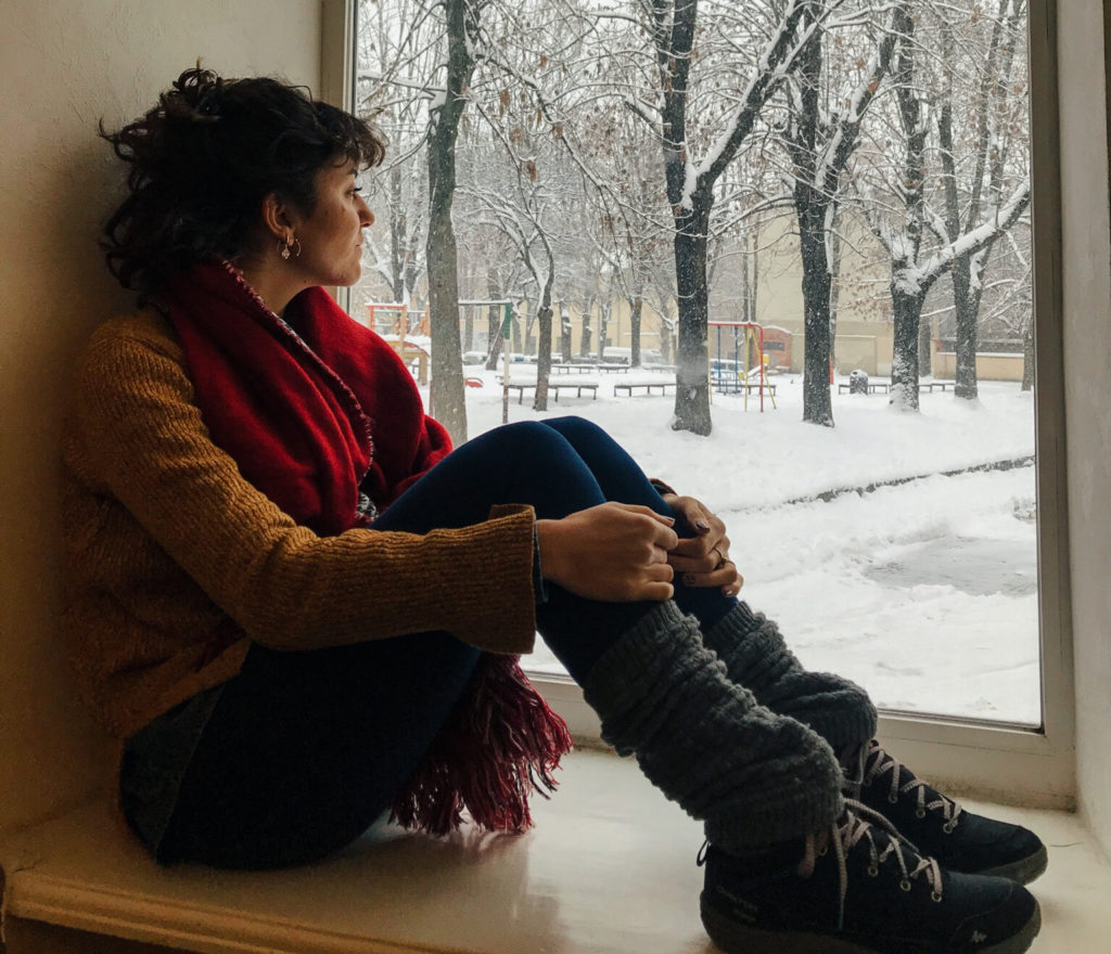 Pencerenin önünde oturup, karla kaplı sokağa doğru bakar kadının çekilmiş görüntüsü.