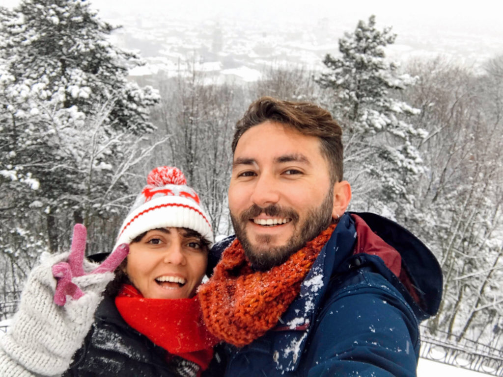 Karla kaplı ağaçların yer aldığı bir parkta, gülümseyip poz veren çiftin mutlu görüntüsü.