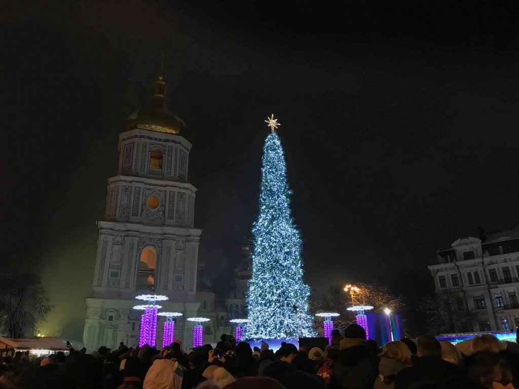 Akşam karanlıkta toplanmış insanlar, ışıklarla aydınlatılmış katedral, yeni yıl ağacı yer aldığı fotoğraf karesi.