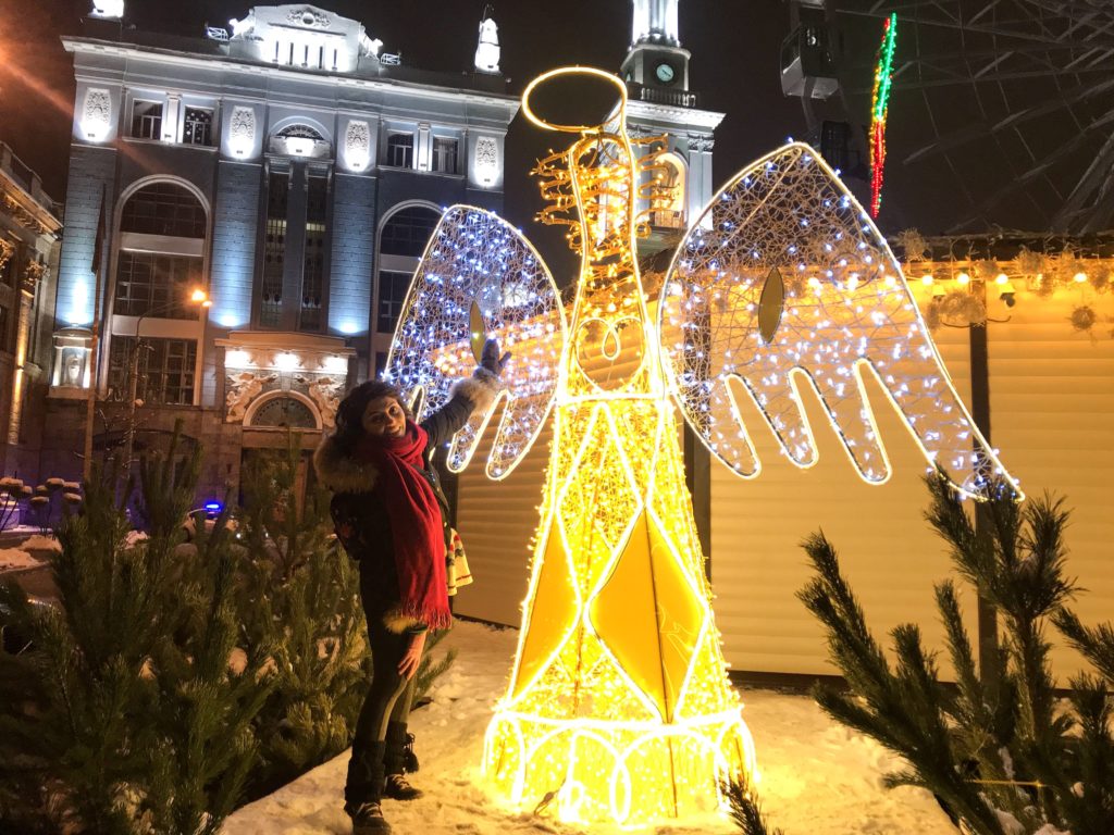 Sarı beyaz ışıklarla süslenmiş melek şeklindeki objenin önünde kanadına elini koyup poz veren GezginStar'ın Kiev yılbaşı yazısından bir görüntüsü.