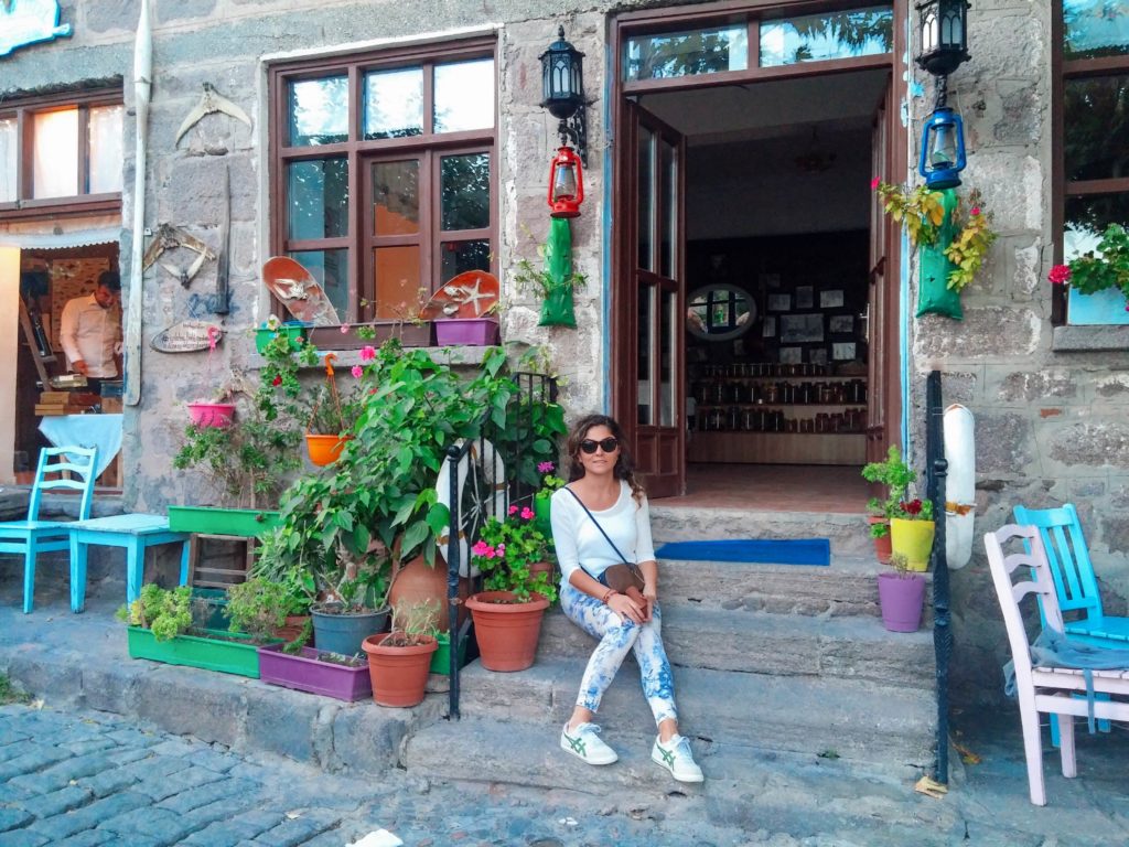 Bir işletmenin kapısının önündeki merdiven basamağına oturup, yanında çiçekler ile poz veren kadının görüntüsü.